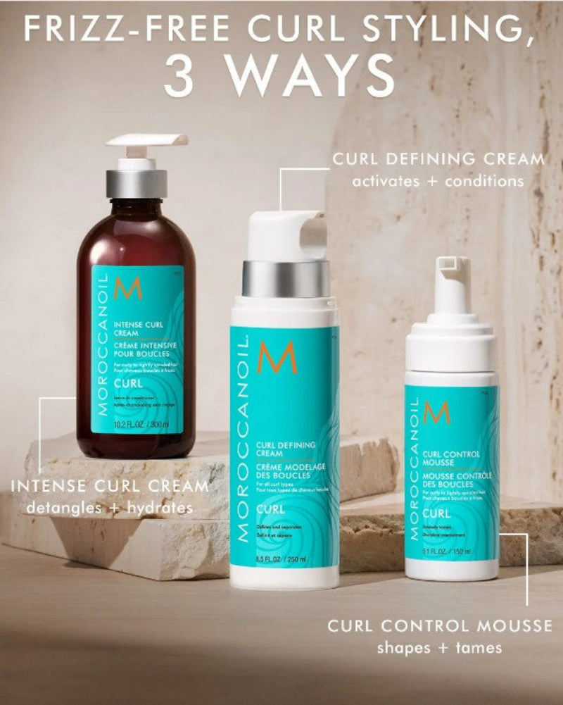 Three Moroccanoil curl styling products displayed on a stone surface with promotional text overlays: an amber bottle of Intense Curl Cream, a white pump bottle of Curl Defining Cream, and a white pump bottle of Curl Control Mousse.