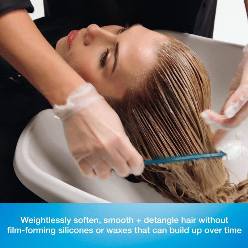 A woman with wet brown hair sits in a salon-style white sink while another person wearing clear gloves applies a smoothing product. A blue comb is used to detangle the hair, and a blue text banner at the bottom reads: 'Weightlessly soften, smooth + detangle hair without film-forming silicones or waxes that can build up over time.