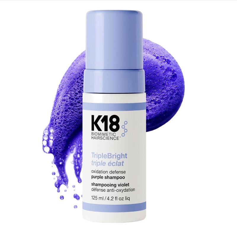 A bottle of K18 TripleBright Triple éclat purple shampoo, featuring a white bottle with a light blue cap, set against a vibrant purple shampoo foam splash on a white background.