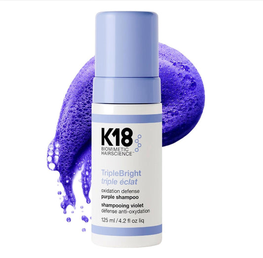 A bottle of K18 TripleBright Triple éclat purple shampoo, featuring a white bottle with a light blue cap, set against a vibrant purple shampoo foam splash on a white background.