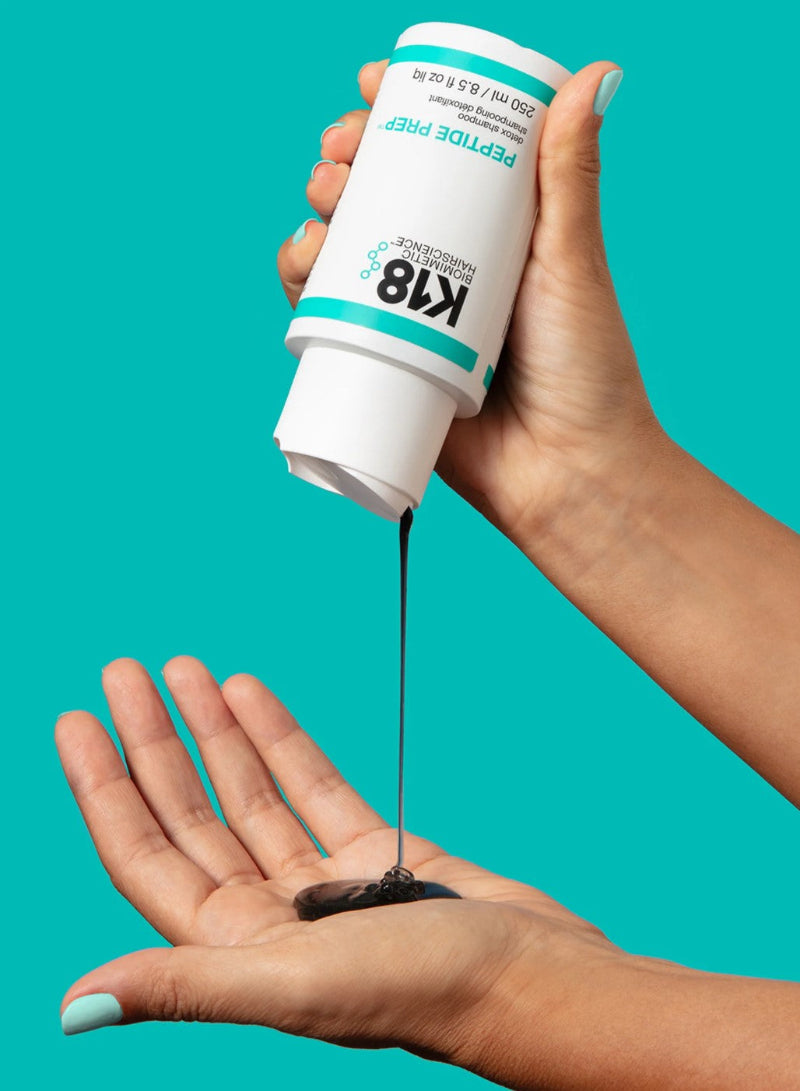 A hand holds a white bottle of K18 Peptide Prep serum, pouring a dark gel into the palm of another hand against a solid teal background.