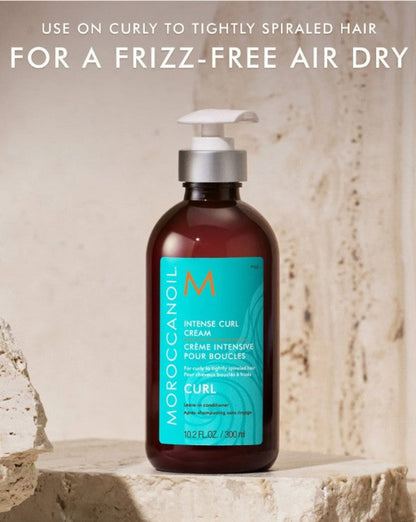 Moroccanoil Intense Curl Cream bottle for a frizz-free air dry hair in an amber brown glass container with a turquoise label and white pump, displayed on a stone surface with promotional text above.