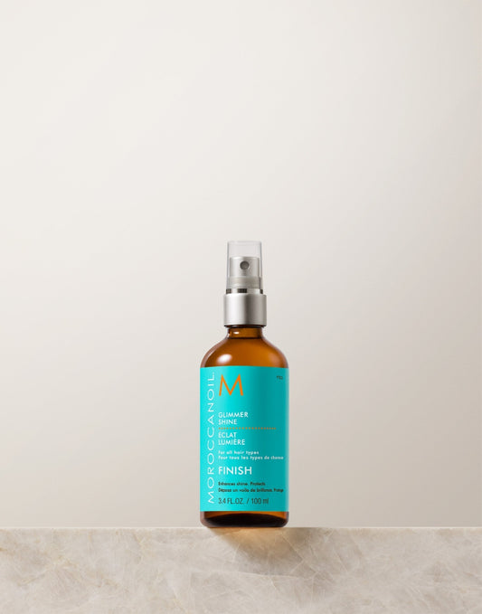 Bottle of hair product with a blue label on a light background. This bottle belongs to Moroccanoil glimmer shine oil for all hair types