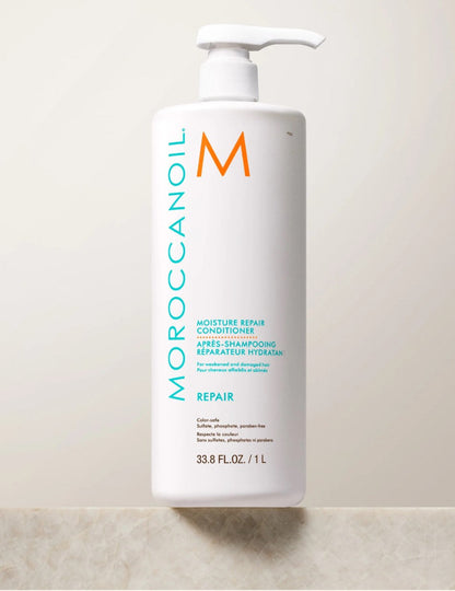 A white bottle of Moroccanoil Moisture Repair Conditioner  1 liter with a pump dispenser, standing on a light beige stone surface against a matching beige background.