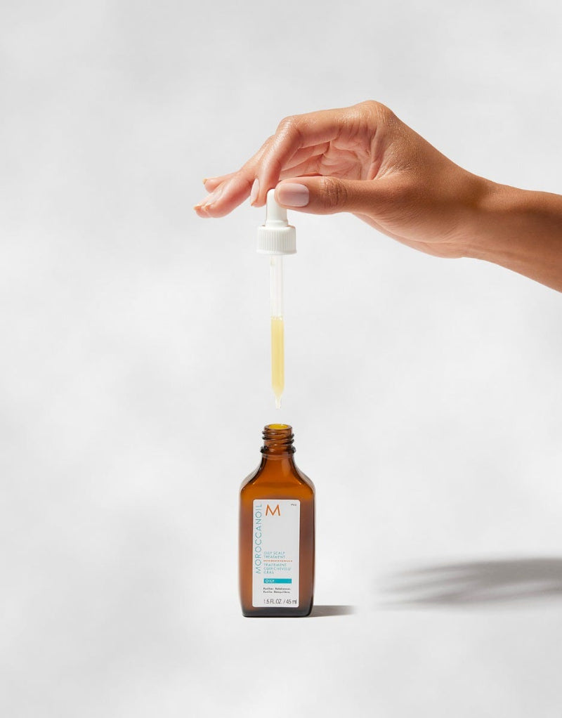Hand holding a dropper over a brown bottle of Moroccanoil oily scalp treatment with a white background