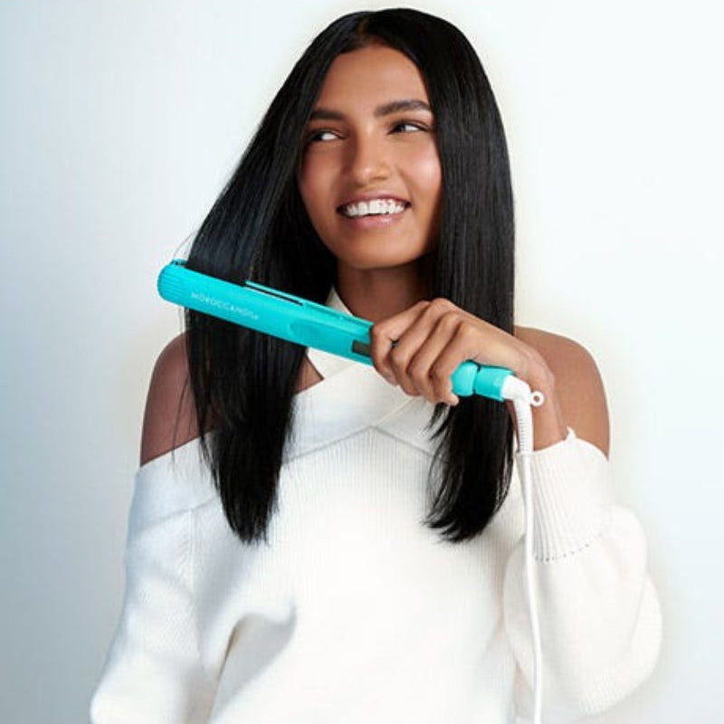 Woman using a Moroccanoil teal colour hair straightener on her black hair against a white background