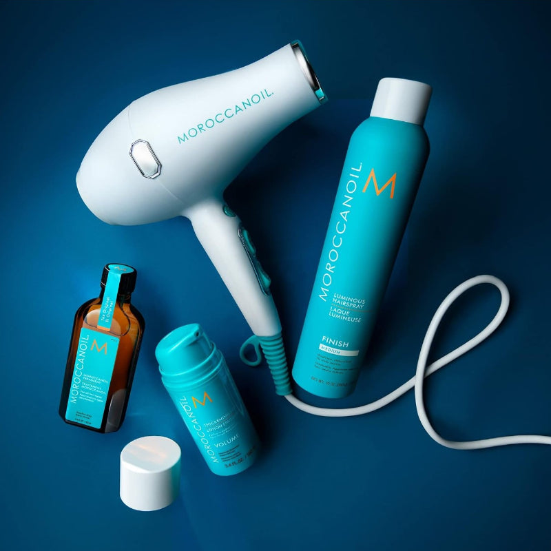 Moroccanoil hair care set arranged on a dark blue background, including a white hair dryer, a turquoise styling foam can and other products for the best hair volume result
