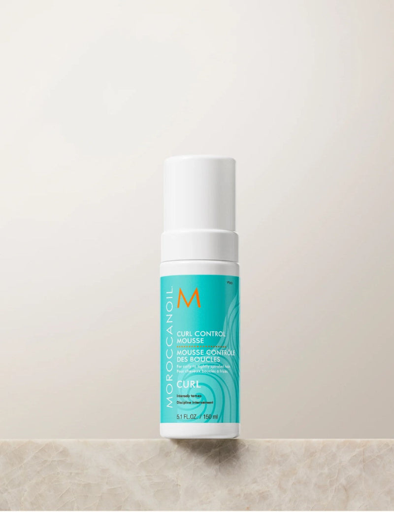 Moroccanoil Curl Control Mousse in a white cylindrical bottle with a turquoise label, standing on a light beige stone surface against a matching beige background.