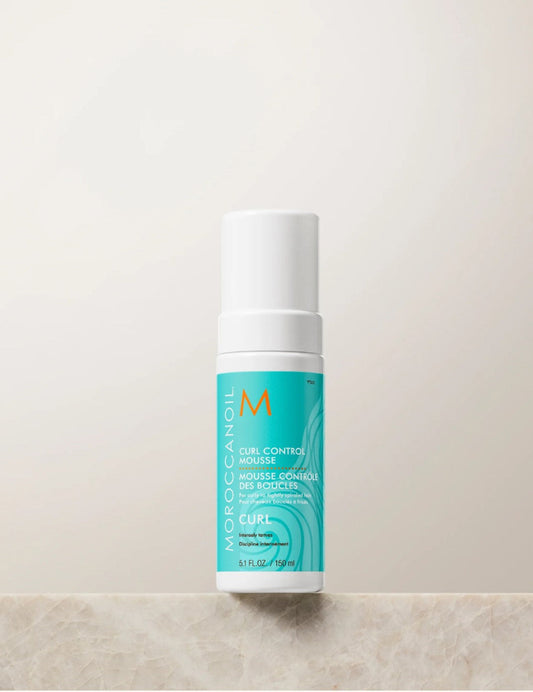 Moroccanoil Curl Control Mousse in a white cylindrical bottle with a turquoise label, standing on a light beige stone surface against a matching beige background.