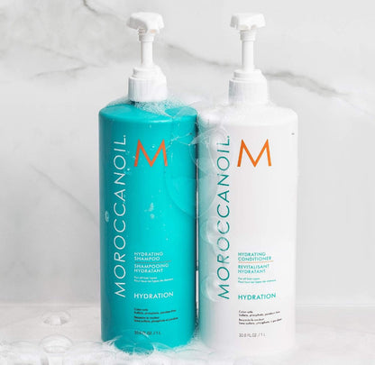 TwoMorocanoil Hydration shampoo and conditioner bottles on a white marble surface with soap bubbles. The shampoo bottle is turquoise and the conditioner bottle is white, both with pump dispensers.