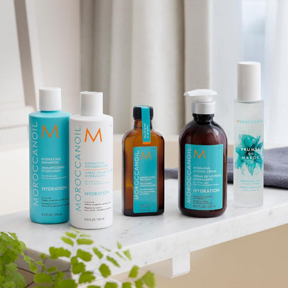 Five Moroccanoil hydration skincare bottles arranged on a white windowsill with soft natural light, a curtain behind, and a small plant in the foreground. The set includes a turquoise bottle, a white bottle, an amber bottle with a turquoise label, a larger amber pump bottle with a turquoise label, and a frosted bottle with a turquoise floral label.