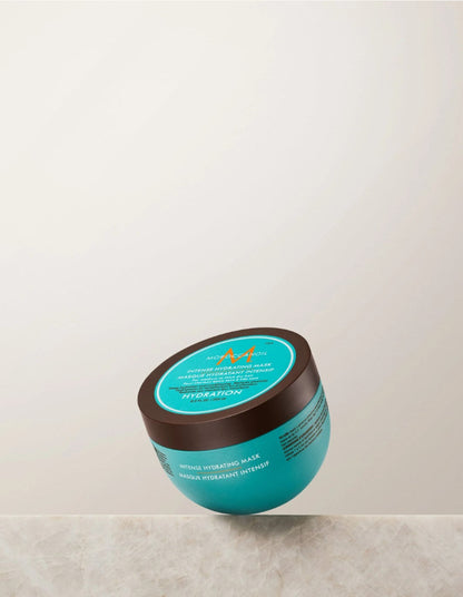 A teal and brown jar of  Moroccanoil Hydration Intense Mask resting on a light marble surface against a neutral off-white background.