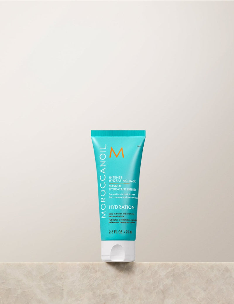 Moroccanoil Morocco Intensive Hydrating Mask in a turquoise squeeze tube travel size with a white cap, standing on a light beige stone surface against a matching beige background.