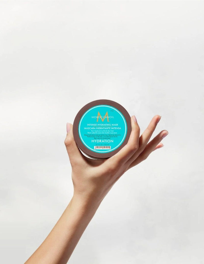 A hand holding a round jar of Moroccanoil Hydration Mask against a clean, light gray background.