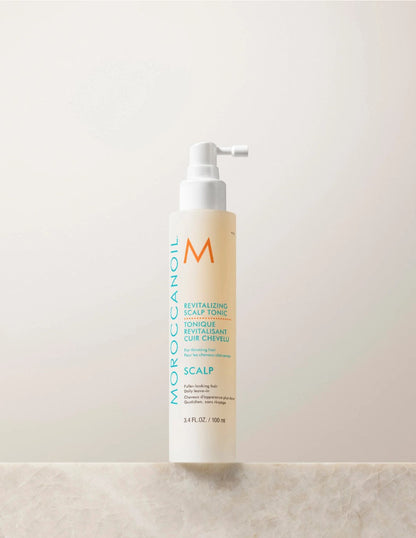 A bottle of Moroccanoil revitalizing scalp tonic for thinning hair on a beige background