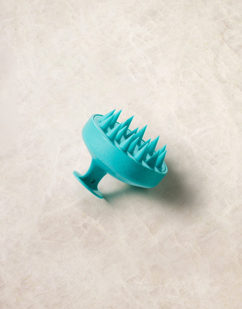 Turquoise Moroccanoil silicone scalp massager on a textured beige surface