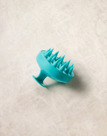 Turquoise Moroccanoil silicone scalp massager on a textured beige surface