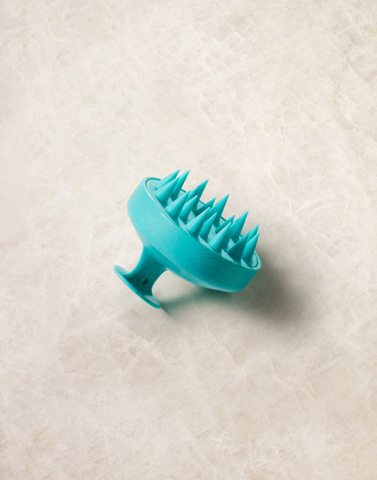 Turquoise Moroccanoil silicone scalp massager on a textured beige surface