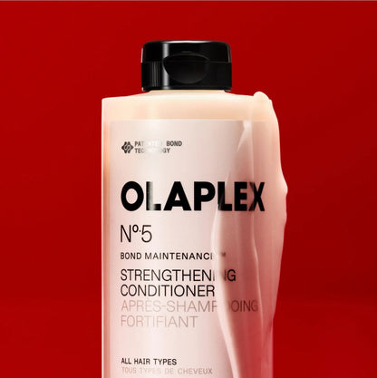 A bottle of Olaplex No. 5 Bond Maintenance Strengthening Conditioner, featuring a light pink liquid visible through the translucent bottle and a black cap, set against a solid red background.