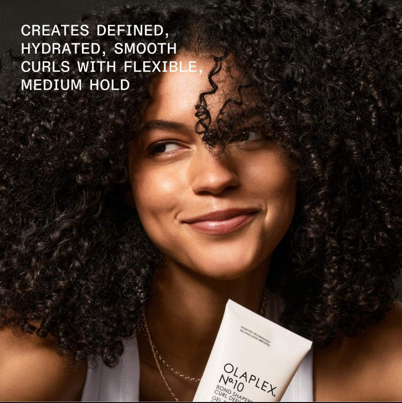 A woman with defined, hydrated, and smooth medium-hold curls is shown in a close-up portrait, holding a white tube of Olaplex No.10 Hair Building Treatment.