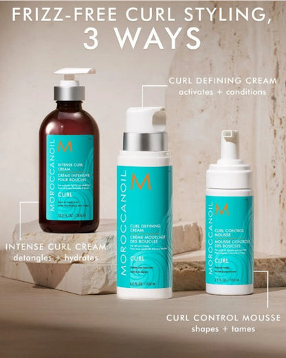 Three Moroccanoil curl styling products displayed on a stone surface with promotional text overlays: an amber bottle of Intense Curl Cream, a white pump bottle of Curl Defining Cream, and a white pump bottle of Curl Control Mousse.