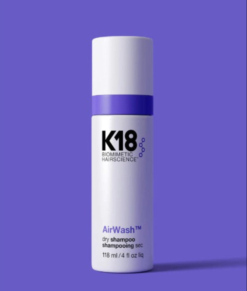 A white cylindrical bottle of K18 Cosmetics AirWash dry shampoo, featuring a purple band and purple text, set against a solid purple background.