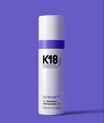 A white cylindrical bottle of K18 Cosmetics AirWash dry shampoo, featuring a purple band and purple text, set against a solid purple background.
