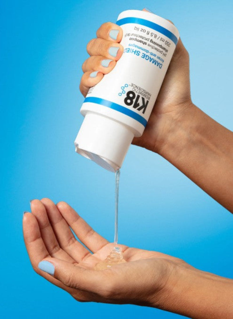 A person's hands holding a white bottle of K18 Damage Shield restoration liquid, with the clear gel being poured from the spout into an open palm against a bright blue gradient background.