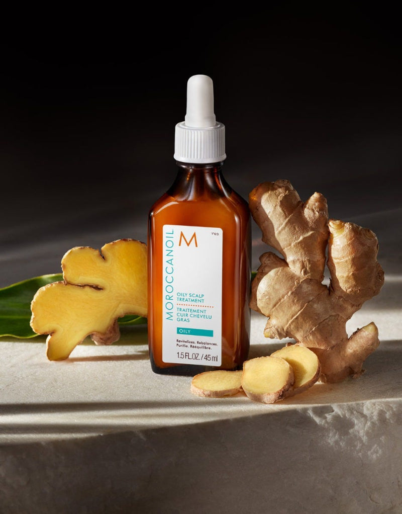 Brown bottle of Moroccanoil oily scalp treatement oil with white cap labeled 'Moroccanoil' on a dark background with ginger root and leaves.