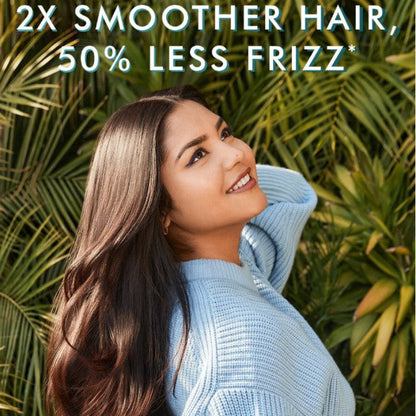A woman with long, straight, glossy brown hair smiles outdoors in front of green palm leaves, wearing a light blue knit sweater. Large text at the top reads, '2X SMOOTHER HAIR, 50% LESS FRIZZ'.